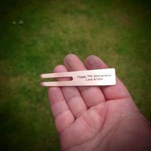 May include: A copper-colored golf divot repair tool with a two-pronged fork design. The tool has the inscription "Happy 7th Anniversary Love Krista". The tool is held in a hand against a green grass background.