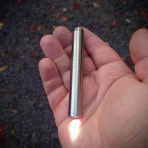 Może przedstawiać: Srebrna metalowa latarka z napisem "FUTURE RETRO GPU" wygrawerowanym z boku. Cylindryczna latarka emituje jasne światło od dołu. Latarka jest trzymana w dłoni osoby.