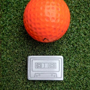 May include: A silver cassette tape charm with a star design on a green artificial grass background. A bright orange golf ball is above the charm.