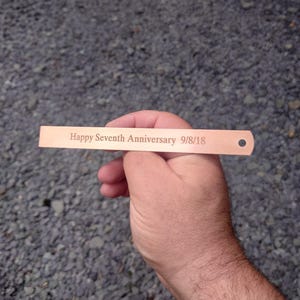 Può includere: Un'etichetta rettangolare marrone chiaro con bordi arrotondati e un piccolo foro. L'etichetta è incisa con le parole "Happy Seventh Anniversary 9/8/18". L'etichetta è tenuta nella mano di una persona su uno sfondo di ghiaia grigia.
