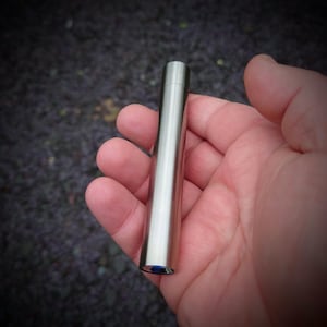 Op de afbeelding: Een cilindrisch object van geborsteld metaal met een afgeronde bovenkant en een blauwe basis wordt in een hand gehouden. Het object heeft een glad, reflecterend oppervlak met een gepolijste afwerking. De achtergrond is wazig, met een donker, getextureerd oppervlak.