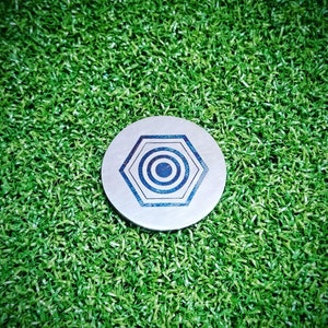 May include: A silver metal disc with a blue and white concentric circle design inside a hexagon. The disc is on a green artificial grass surface.