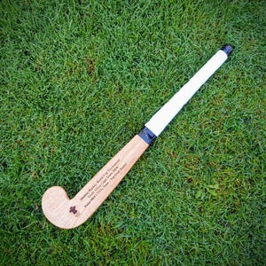 Puede incluir: Un palo de hockey sobre hierba de madera con un agarre blanco y cinta negra, sobre hierba verde. El palo tiene texto que dice: "Masters Hockey World Cup Tournament South Africa Cape Town 2004 Wales Men's O35s Team - Top Goal Scorer."