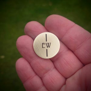 Può includere: Un piccolo disco metallico circolare color oro spazzolato con una linea verticale che attraversa il centro e le lettere maiuscole E e W incise su entrambi i lati della linea. La superficie ha una finitura opaca e un design piatto.