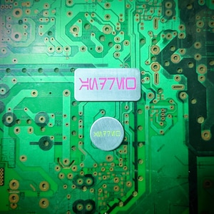 May include: Close-up of a green circuit board with copper-colored traces and components. Two metallic labels are affixed to the board, one rectangular and one circular, each bearing the text "VK77IVO" in a stylized font.