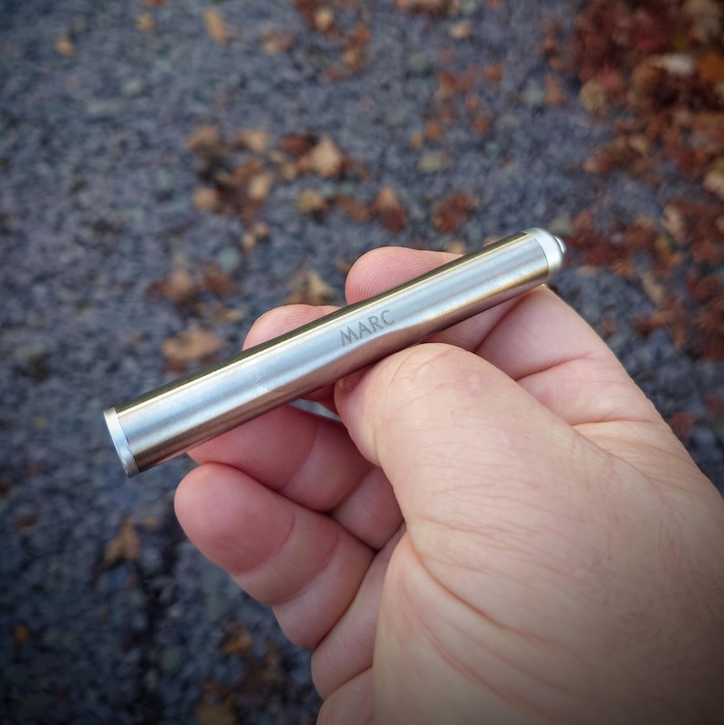 Pu&ograve; includere: Un oggetto cilindrico in metallo argentato con il nome "MARC" inciso sul lato. L'oggetto &egrave; tenuto in mano su uno sfondo sfocato di foglie e piccole rocce. L'oggetto ha un'estremit&agrave; arrotondata e un'estremit&agrave; piatta.