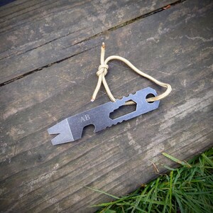 Könnte beinhalten: Ein silbernes Multitool mit einem Flaschenöffner, einem Brecheisen und anderen Werkzeugen. Das Werkzeug ist mit den Buchstaben "AB" graviert und an einem beigefarbenen Paracord befestigt.
