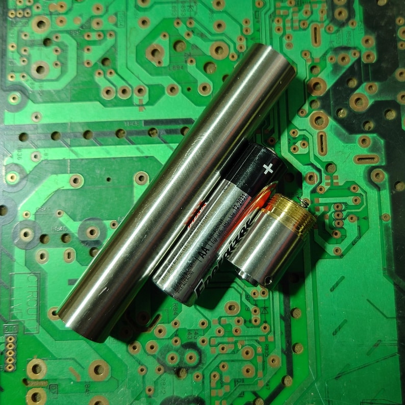 Pu&ograve; includere: Un assortimento di componenti elettronici, tra cui un tubo di metallo argentato, un cilindro filettato color oro e due batterie AA. I componenti sono disposti su un circuito stampato verde.