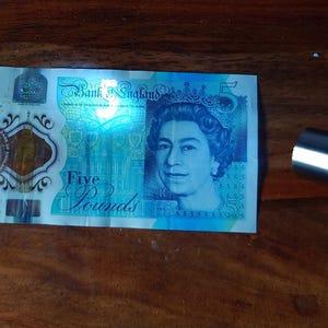 Pu&ograve; includere: Una banconota da cinque sterline della Banca d'Inghilterra, con un ritratto della regina Elisabetta II. La banconota &egrave; prevalentemente blu e verde, con il numero "5" ben visibile. Un oggetto argentato &egrave; sullo sfondo.