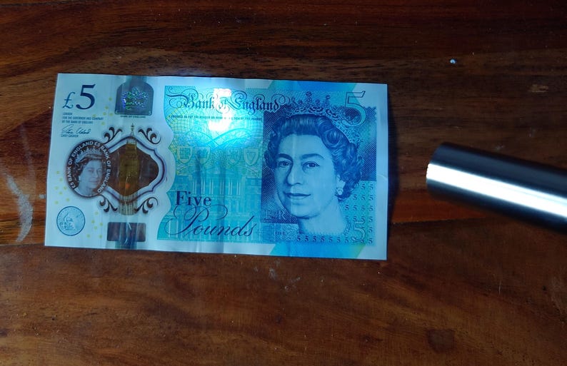 Pu&ograve; includere: Una banconota da cinque sterline della Banca d'Inghilterra, con un ritratto della regina Elisabetta II. La banconota &egrave; prevalentemente blu e verde, con il numero "5" ben visibile. Un oggetto argentato &egrave; sullo sfondo.