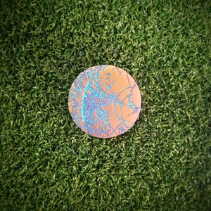 Puede incluir: Un disco redondo de color cobre con un diseño abstracto azul y blanco. El disco está sobre una superficie verde.