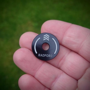 May include: Black round disc with a white circle around the edge and the word "RADFORD" printed in white. The center of the disc has a small hole and three white arrows pointing upwards.