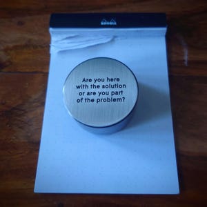 Pode incluir: Um peso de papel circular em metal prateado escovado com o texto preto gravado: Are you here with the solution or are you part of the problem? O objeto está posicionado sobre um bloco de notas Rhodia branco com encadernação superior preta.