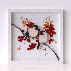 Puede incluir: Una pieza de arte de pared de corte por láser enmarcada que presenta dos pájaros posados en una rama con bayas rojas. Los pájaros son blancos con detalles negros y la rama es marrón. El fondo es una textura de madera blanca.