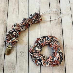 May include: A set of two hair scrunchies with a floral print in shades of pink, orange, and black on a white background. One scrunchie is attached to a white elastic cord with a silver clip.
