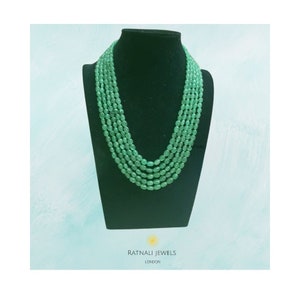 Könnte beinhalten: Eine mehrsträngige Halskette mit vier Reihen glatter, oval geformter grüner Perlen. Die Kette wird auf einer schwarzen Büste präsentiert. Unter der Büste steht der Text RATNALI JEWELS LONDON mit einem kleinen gelben sonnenartigen Symbol.