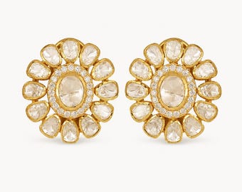 May include: A pair of gold stud earrings featuring a central oval-shaped stone surrounded by a halo of small round stones, with an outer ring of larger, petal-shaped stones. The design creates a floral appearance with clear, faceted stones.