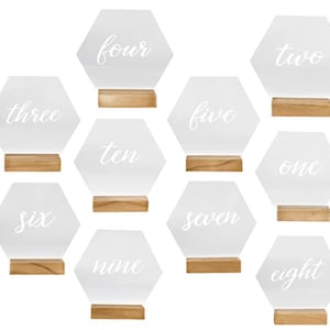 Op de afbeelding: Tien transparante, zeshoekige tafelnummerborden van acryl met witte kalligrafie-nummers van één tot tien. Elk bord heeft een kleine houten standaard.