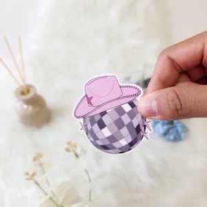 May include: A pink cowboy hat with a star on top sits on top of a silver disco ball with white squares. The disco ball has two silver stars on either side.