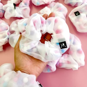 May include: White mesh hair scrunchies with colorful pom poms. The scrunchies have a black label with the letters "JR" on them.