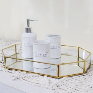 Op de afbeelding: Een gouden, achthoekige, gespiegelde dienblad met drie witte keramische containers. De containers hebben de woorden "Wash", "Water" en "Brush" erop geschreven. Het dienblad staat op een wit gehaakt oppervlak.