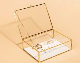 May include: A rectangular glass jewelry box with a gold-colored metal frame and a hinged lid. Inside, the box contains various jewelry items, including a gold-colored hoop earring, a pearl hair clip, and a pair of earrings with a geometric design. The box is open, revealing the contents.
