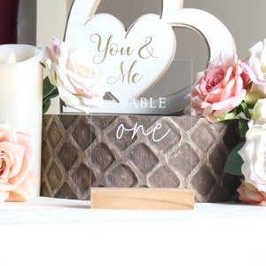 May include: Clear acrylic table number sign with the words "Table One" in white script. The sign is on a wooden base and is sitting on a brown wooden surface with a white heart-shaped wooden sign that says "You & Me" in gold script.