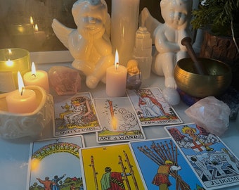 Può includere: Una lettura di tarocchi con sette carte disposte su un tavolo. Le carte sono: Regina di Coppe, Asso di Bacchette, Re di Spade, Otto di Bacchette, Sette di Bacchette, Giudizio e Il Mondo. Ci sono anche candele, cristalli e figurine sul tavolo.