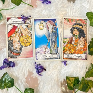May include: Three tarot cards, The Hermit, Queen of Pentacles, and Page of Pentacles, with colorful illustrations and text in multiple languages.