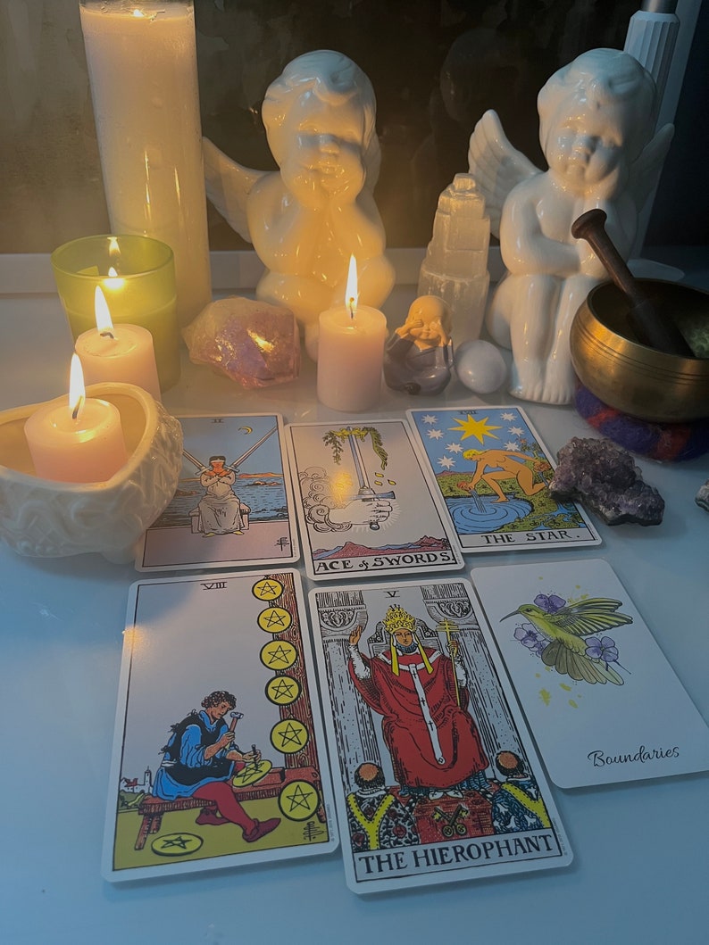 Pu&ograve; includere: Una lettura di tarocchi con uno spargimento di otto carte, tra cui Il Gerofante, La Stella, Asso di Spade, Cinque di Pentacoli e una carta con un colibr&igrave;. Le carte sono disposte su una superficie bianca con due statuine di angeli bianche, candele e cristalli.