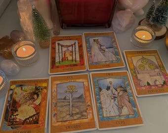 Può includere: Carte dei tarocchi disposte su un tavolo con una candela accesa sullo sfondo. Le carte mostrano immagini di una regina, un due di coppe, un tre di pentacoli, un asso di spade, un tredici di bastoni e un tredici di coppe.