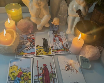 Può includere: Una scena spirituale con candele accese, statuette di angeli, cristalli e carte dei tarocchi. Le carte includono "Knight of Swords", "King of Swords" e "Abundance". Sono presenti anche una piccola ciotola tibetana e una statuina di monaco.
