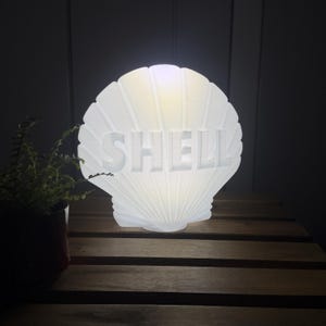 May include: A white, seashell-shaped lamp with the word "SHELL" cut out in the center. The lamp is lit from within, creating a soft glow. The lamp is sitting on a wooden surface.