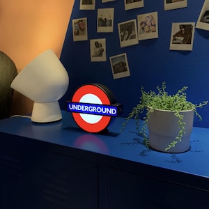 May include: A circular illuminated sign featuring the iconic red ring and blue horizontal bar with the white text UNDERGROUND, positioned next to a white modern table lamp and a potted succulent plant on a blue surface.