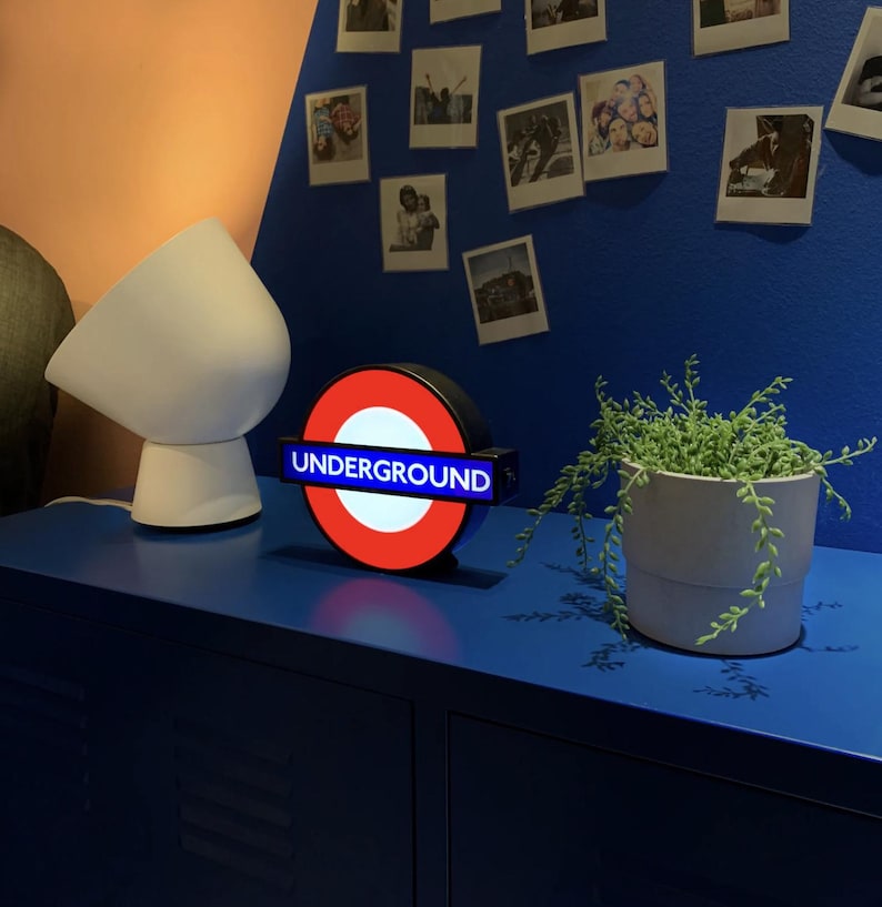 May include: A circular illuminated sign featuring the iconic red ring and blue horizontal bar with the white text UNDERGROUND, positioned next to a white modern table lamp and a potted succulent plant on a blue surface.