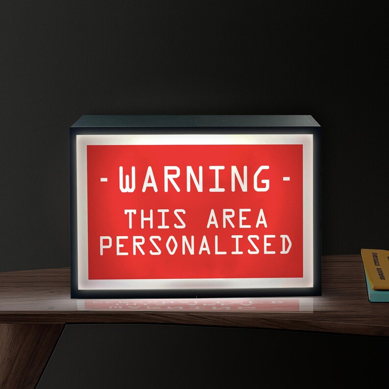 May include: A black rectangular light box with a red illuminated panel. The panel displays the white text: "- WARNING - THIS AREA PERSONALISED". The light box is on a wooden surface.