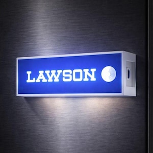 Puede incluir: Un letrero rectangular iluminado con la palabra "LAWSON" en letras mayúsculas blancas sobre un fondo azul. Un círculo blanco está a la derecha del texto. El letrero está enmarcado en blanco y montado en una pared gris oscuro.