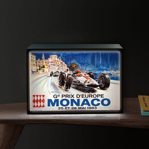 May include: A framed vintage poster featuring a race car speeding along a coastal road. The poster has a blue and white color scheme with the text "Gd PRIX D'EUROPE MONACO 25 ET 26 MAI 1963". The poster is in a black frame.