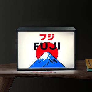 May include: A rectangular light box with a graphic of Mount Fuji. The design features a red sun, a blue and white mountain, and the word "FUJI" in black and red. The light box is on a wooden surface.