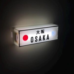 Puede incluir: Un letrero rectangular iluminado con la palabra "OSAKA" en letras negras. El letrero también presenta un círculo rojo y un círculo azul, con caracteres japoneses encima del texto en inglés. El letrero es blanco con un borde negro.