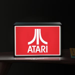 May include: A rectangular light box featuring a vibrant red background with the white Atari logo and the word ATARI printed in white capital letters. The sign is encased in a black frame and emits a bright glow from the front display panel.