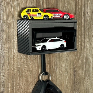 Peut inclure: Un porte-clés mural noir avec un design de garage miniature. Le garage contient une petite voiture blanche, et deux autres voitures miniatures sont garées au-dessus. Le porte-clés a un motif en fibre de carbone sur le côté et un crochet pour les clés.