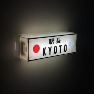 May include: A rectangular, illuminated sign with the word "KYOTO" in black capital letters. Above the word is Japanese text. A red circle and a blue circle are on the sign. The sign is white with a black border.