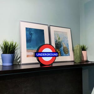 May include: A circular London Underground sign featuring a red ring, a white center, and a blue horizontal bar with the word UNDERGROUND in white capital letters. The sign is illuminated and sits on a dark shelf between two framed art prints.