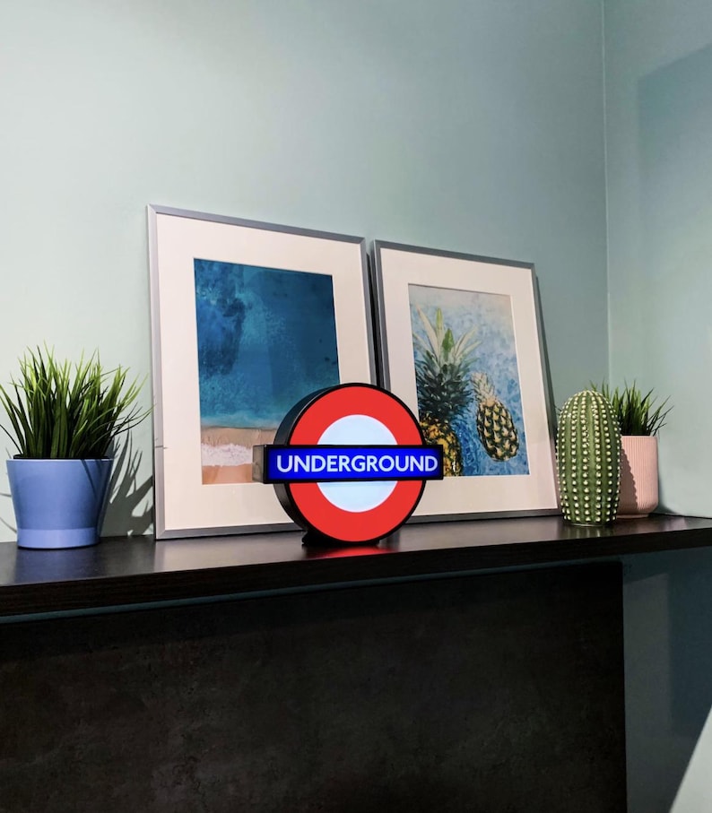 May include: A circular London Underground sign featuring a red ring, a white center, and a blue horizontal bar with the word UNDERGROUND in white capital letters. The sign is illuminated and sits on a dark shelf between two framed art prints.