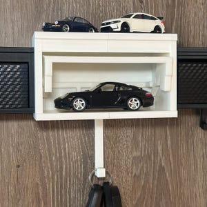 Könnte beinhalten: Ein weißer, an der Wand befestigter Schlüsselhalter mit einem Regal, auf dem drei Spielzeugautos ausgestellt sind: ein schwarzer Porsche, ein dunkelblaues Auto und ein weißes Auto. Der Schlüsselhalter hat einen weißen Haken für Schlüssel und schwarze Seitenteile.