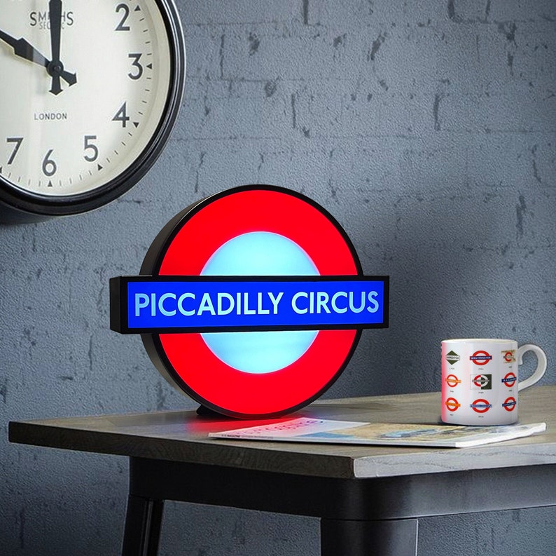 Peut inclure: Un panneau lumineux rouge et bleu du m&eacute;tro londonien avec les mots "Piccadilly Circus" dessus. Le panneau est &eacute;clair&eacute; et a un cercle blanc au centre.