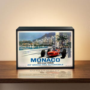May include: A framed light box featuring a vintage poster for the Monaco Grand Prix. The poster depicts a red race car on a track with the city in the background. Text includes "MONACO" and "30 mai 1965".