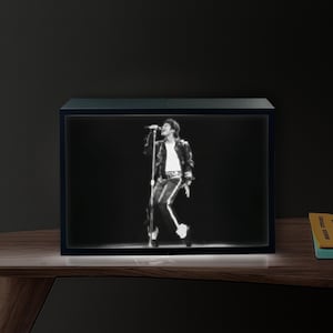 May include: A black and white framed image of a singer performing on stage. The singer is wearing a black jacket, white shirt, and dark trousers. The frame is rectangular and sits on a wooden surface.