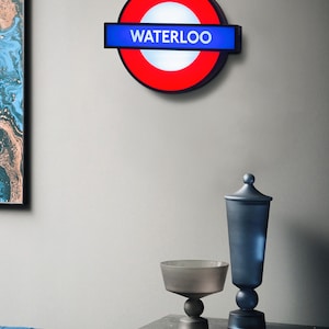 Peut inclure: Un panneau de m&eacute;tro londonien rouge et bleu avec le mot "WATERLOO" dessus. Le panneau est mont&eacute; sur un mur blanc.